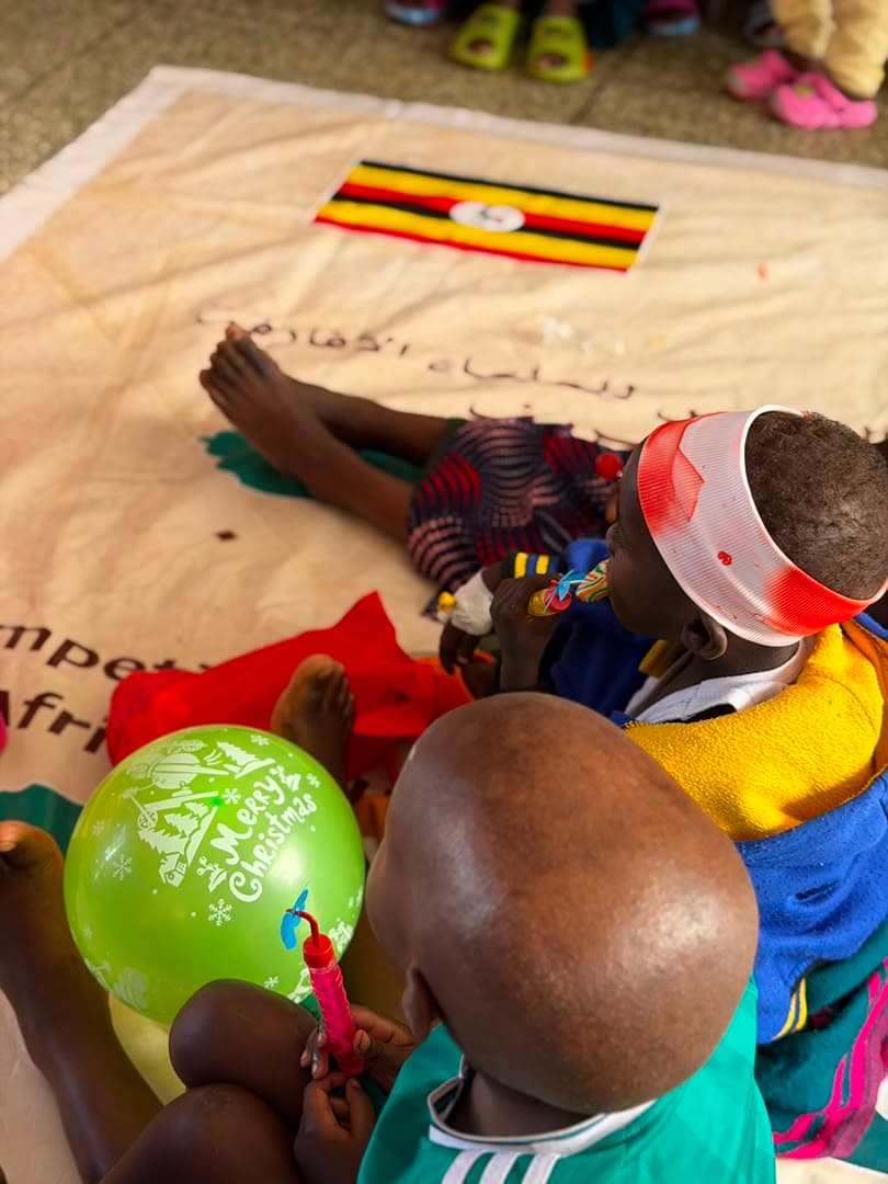 Children sitting around a signed banner printed with the Ugandan flag, one holding a green balloon