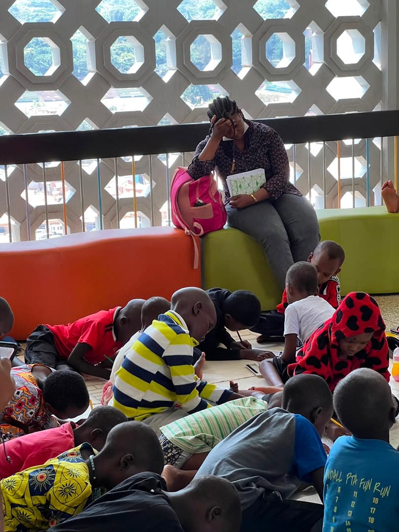 A volunteer sitting at the front of the ward reading a picture book aloud to children gathered on the floor